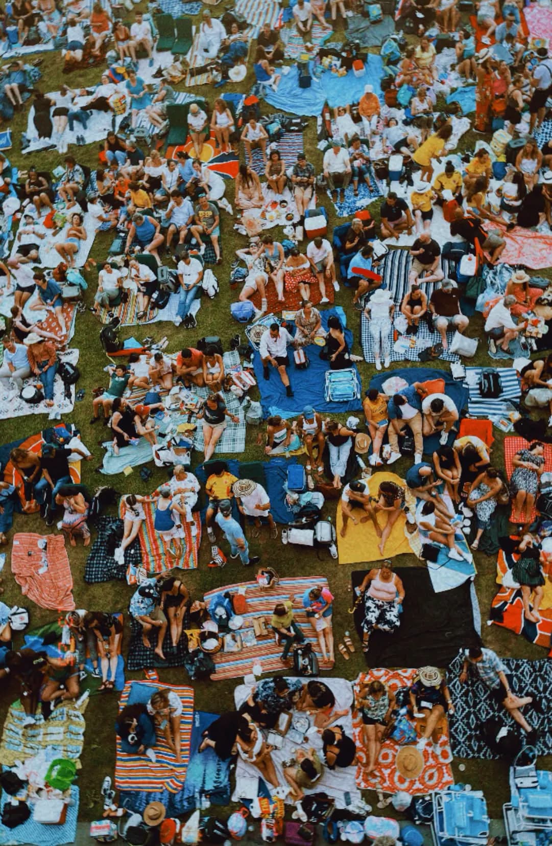 A large public outdoor gathering viewed from above, with people seated together across blankets in a shared community setting.