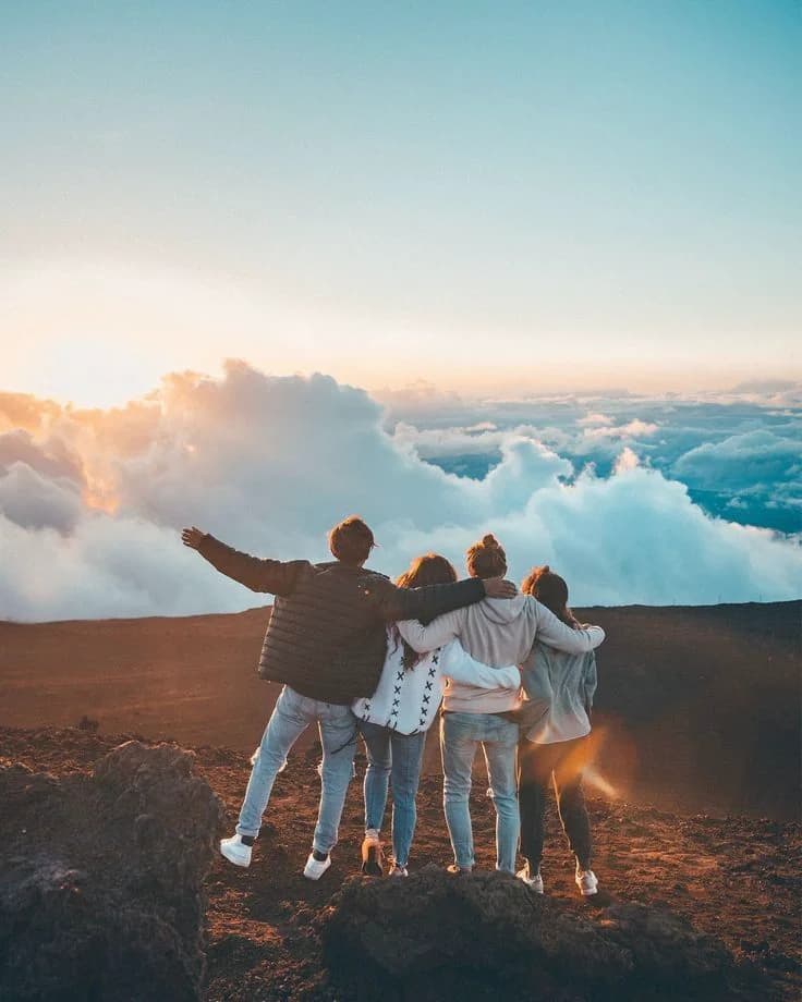 Four friends standing together on a mountaintop at sunrise, looking across the clouds.